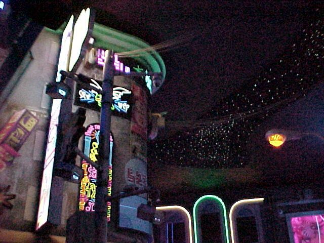 A low res photo taken in the interior of Meow Wolf Convergence Station in Denver, CO. It resembles a futuristic city scene with glowing neon signs on a building, traffic lights, wires, and stars above.